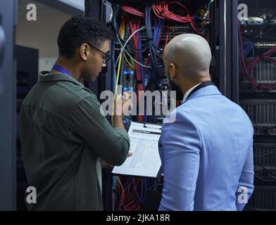 Explaining the ins and outs. two male IT support agents working together in a dark network server room. Stock Photo