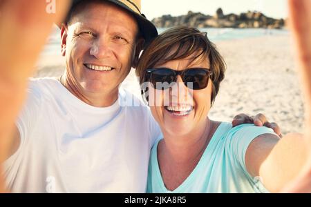 We enjoying our vacation and want to share it with our family. a mature couple taking a selfie while spending the day at the beach. Stock Photo