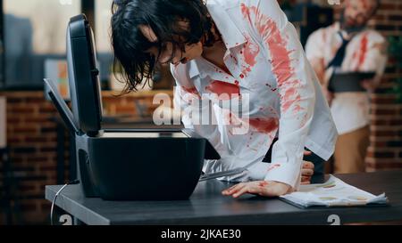 Dumb creepy looking zombie trying to use photocopy machine. Spooky evil brainless monster with deep and bloody scars and wounds while damaging scanner machine in company office workspace. Stock Photo