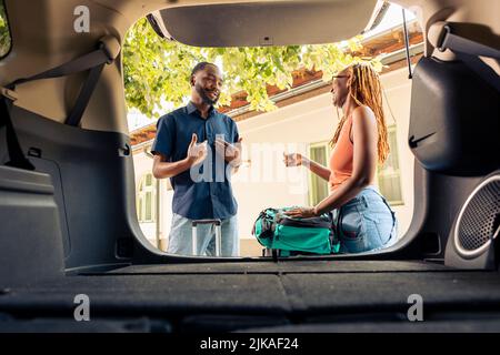 African american couple putting travel bags and trolley in trunk, getting ready to leave on holiday vacation with automobile. Travelling by car on road trip with luggage and suitcases. Stock Photo