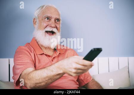 Excited senior man with white beard using remote control to switch channels Stock Photo