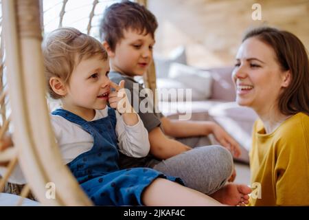 Young cheerful mother playing with her little children and having fun when swinging them on hanging chair in conservatory at home. Stock Photo