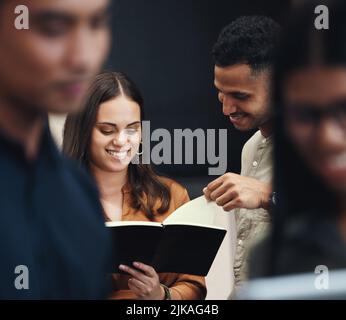 Lets see what youve got there. two young businesspeople looking over a notebook near the back of their office with colleagues in the foreground. Stock Photo
