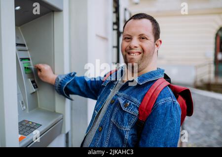 Happy young man with Down sydrome using a street ATM machine and withdrawing money. Stock Photo