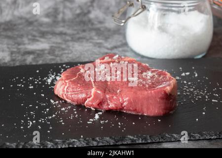 Rump beef steak, seasoned with sea salt with jar in background, on a slate chopping board. On a dark stone background Stock Photo