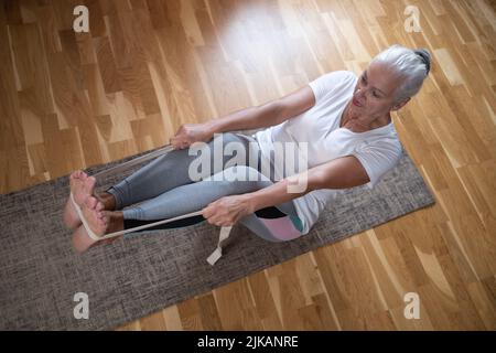 Senior caucasian woman doing abs exercises, sitting in boat yoga pose at home. Stock Photo
