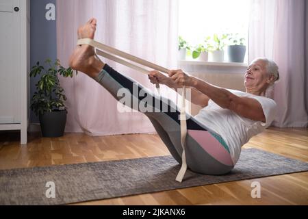 Senior caucasian woman doing abs exercises, sitting in boat yoga pose at home. Stock Photo