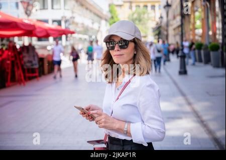 Middle aged woman holding cellphone in her hand and text messaging while walking on the street. Confident woman wearing baseball cap and sunglasses. Stock Photo
