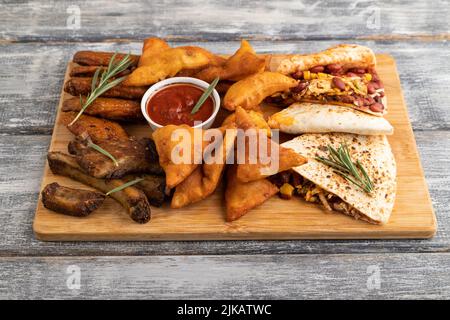 set of snacks: nachos, chicken wings, fried ribs, bean tortillas on a cutting board on a gray wooden background. Side view, close up. Stock Photo