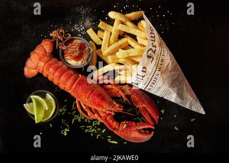 Top view of yummy red lobster and potato chips in newspaper sheet served with sauce and herbs near lime slices on black background Stock Photo