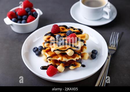 Viennese or Belgian waffles with fresh berries (raspberries and blueberries) on a white plate and a cup of coffee. Traditional dessert. Close-up, grey Stock Photo