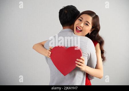 Ecstatic young woman embraced by her boyfriend holding heart-shaped gift box and looking at camera Stock Photo