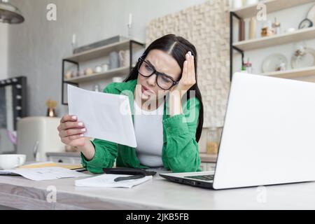 Distressed young woman sit on couch at home read bad negative news in paper correspondence, unhappy biracial female stressed consider unpleasant message in postal letter Stock Photo