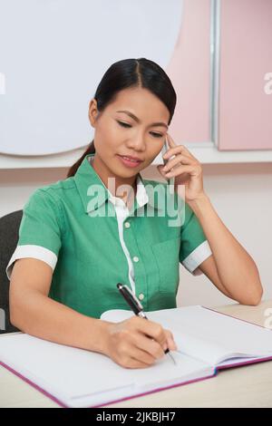 Vietnamese female spa owner talking on the phone with client and taking notes Stock Photo