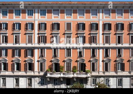 Apartment building with balconies and shuttered windows, some closed, in an orange plastered wall on a sunny day in Rome, Italy. At the top of a borde Stock Photo