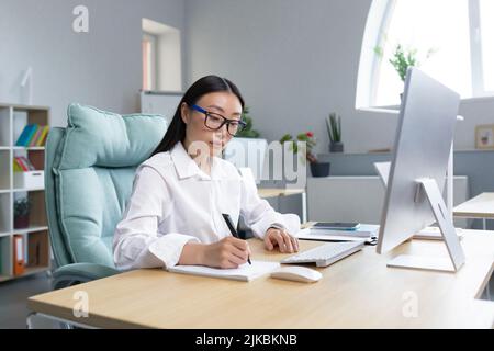 Young beautiful Asian woman secretary making notes in notebook with pen, sitting at desk in office wearing glasses and white shirt. Stock Photo