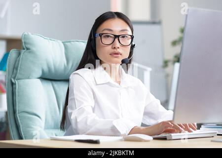 Portrait of a young Asian woman in headphones with a microphone. Works as a consultant in a call center. Sitting at the table in glasses at the computer, looking at the camera, smiling. Stock Photo
