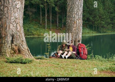 Young Vietnamese couple sitting under the tree and reading something on tablet computer Stock Photo