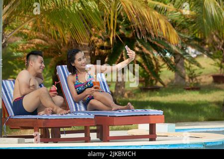 Happy young couple sunbathing by the pool and taking selfies Stock Photo