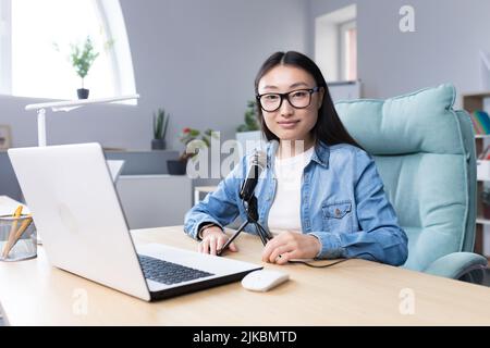 Podcast recording, interview. Young beautiful Asian woman sitting at a table with a microphone and a laptop. Talks, tells. Makes an audio recording of a podcast, gives an interview via video call. Stock Photo