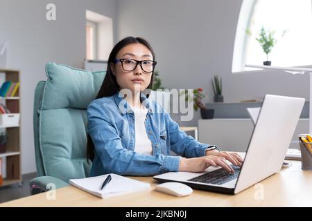 Distance Learning. A young Asian woman is a teacher, conducts online classes, lessons. He looks into the camera, explains, tells. Sitting in glasses and jeans at a table at a computer in a classroom Stock Photo