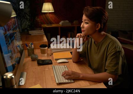 Portrait of female Asian designer looking at images on computer monitor in the light of lamp Stock Photo