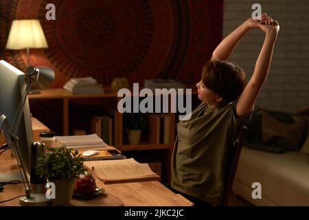 Tired college student stretching while doing homework in dark room in the light of lamp Stock Photo