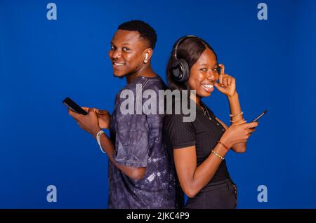 Positive african couple standing back to back, holding and using their mobile phones, chatting online or scrolling social networks feed Stock Photo