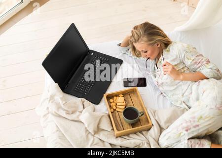 A young woman works while lying in bed - she checks the news in her smartphone. There is a laptop and a breakfast tray on the bed. Freelancer life. Stock Photo