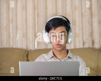 The boy wearing headphones sitting at the table with internet chat skype teacher prepare for exam and using laptop computer for his homework and E-lea Stock Photo