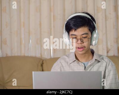 The boy wearing headphones sitting at the table with internet chat skype teacher prepare for exam and using laptop computer for his homework and E-lea Stock Photo