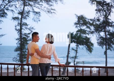Back view of loving Asian couple gently embracing each other while standing on wooden terrace and enjoying breathtaking seascape Stock Photo