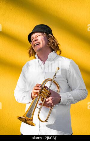 Young caucasian long-haired blond cheerful smiling man with trumpet outdoor Stock Photo