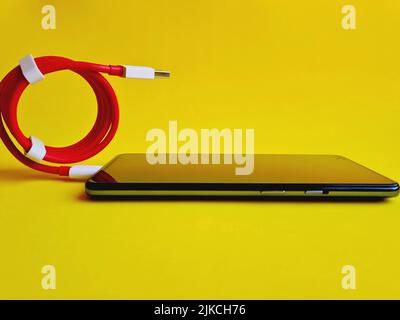 A blue smartphone connected to a new red cable resting on yellow background - Stock Photo