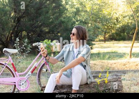 Young man sitting on a tree trunk texting his friends on the smartphone with a pink vintage bike and plants in the background Stock Photo