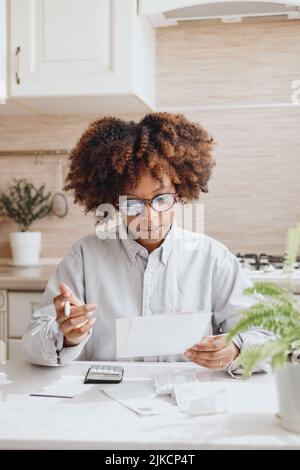 Curly-haired woman pays bills at the kitchen table. Stock Photo
