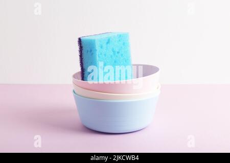 Stack of colourful bowls with blue dishwashing sponge on pink and white background. Close-up. Pastel colours. Cleaning concept. Stock Photo