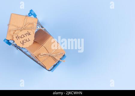 Mini shopping cart on light blue background. Kraft paper boxes with tag inscription Thank you inside trolley. Top view. E-commerce, online shopping Stock Photo