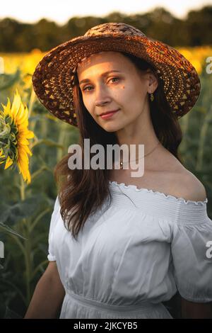 A young girl in a white dress and hat in a field of sunflowers at sunset. Portrait of a woman with a slim figure on a background of yellow flowers Stock Photo