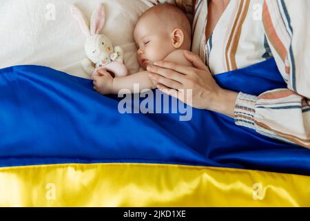 Close-up portrait of a mother kissing her newborn baby sleeping on the bed at home covered with the Blue and yellow flag of Ukraine. The baby sleeps w Stock Photo