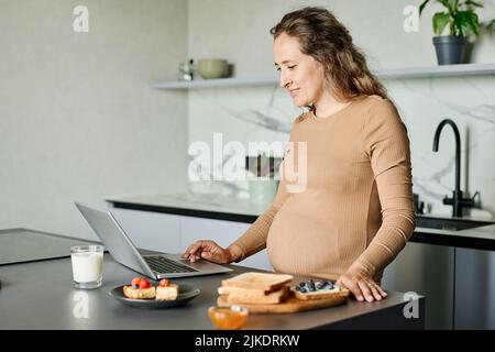Young pregnant woman searching for new recipes of healthy food in the net while standing by kitchen table in front of laptop Stock Photo