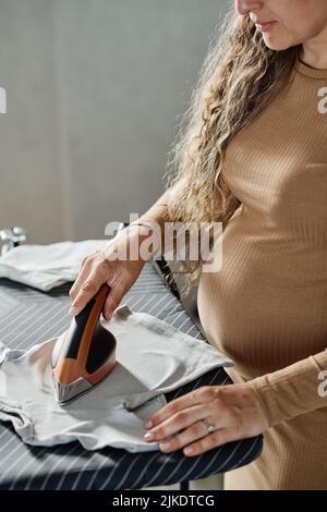 Young pregnant woman in casual dress of beige color ironing clothes of her child with electric iron while standing by board Stock Photo