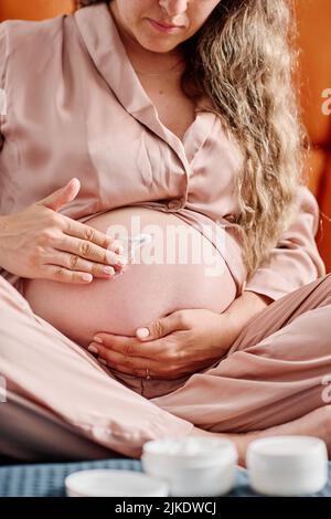 Young pregnant woman in pink pajamas applying nutritive night cream on her belly while taking care of body and skin before sleep Stock Photo