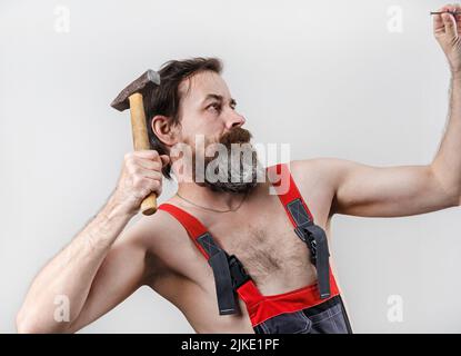 Bearded builder in overalls hammers a nail with a hammer. Man with hammer. Stock Photo