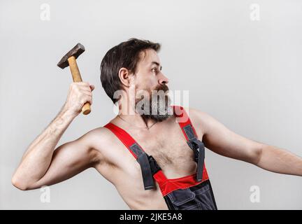 Stern bearded man in overalls swings a hammer. Builder with a hammer. Stock Photo