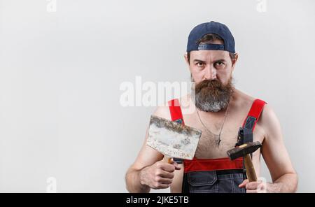 Bearded man in overalls and a baseball cap holds a hammer and spatula. Portrait of a worker. Copy space for your text. Stock Photo