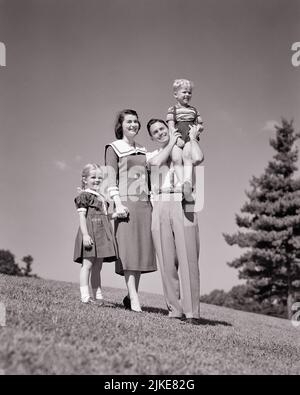 1950s FAMILY ON GRASSY HILLSIDE MOTHER FATHER AND 2 KIDS BOY ON DAD’S SHOULDER GIRL HOLDING HANDS WITH MOM LOOKING OUT AT VIEW - j4275 HAR001 HARS SHOULDER MOM CLOTHING NOSTALGIC PAIR 4 SUBURBAN MOTHERS OLD TIME NOSTALGIA BROTHER OLD FASHION SISTER JUVENILE STYLE PLEASED FAMILIES JOY LIFESTYLE FEMALES MARRIED BROTHERS RURAL SPOUSE HUSBANDS HEALTHINESS HOME LIFE COPY SPACE FRIENDSHIP FULL-LENGTH LADIES PERSONS MALES SIBLINGS SISTERS FATHERS B&W PARTNER SUMMERTIME HAPPINESS CHEERFUL ADVENTURE LEISURE AND DADS EXCITEMENT LOW ANGLE PARKS RECREATION PRIDE SIBLING SMILES CONNECTION NUCLEAR FAMILY Stock Photo