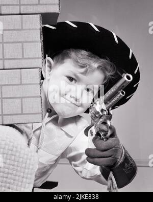 1950s BOY DRESSED AS COWBOY WEARING HAT FRINGED GLOVES HOLDING PISTOL PEEKING AROUND WALL OF CARDBOARD BOXES LOOKING AT CAMERA - j4537 HAR001 HARS WESTERN EXPRESSIONS B&W EYE CONTACT COWBOYS HAPPINESS CARDBOARD HEAD AND SHOULDERS ADVENTURE EXCITEMENT CONCEPTUAL IMAGINATION CAP PISTOL SIX GUN TOY GUN FIREARM FIREARMS FRINGED JUVENILES PISTOL SIX SHOOTER BLACK AND WHITE CAUCASIAN ETHNICITY HAR001 OLD FASHIONED Stock Photo