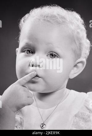 1950s LITTLE BLONDE GIRL TODDLER LOOKING AT THE CAMERA WITH HER INDEX FINGER TO HER LIPS TO INDICATE QUIET SHUSH OR A KISS - j6351 HAR001 HARS COPY SPACE QUIET GESTURING B&W EYE CONTACT INDEX HEAD AND SHOULDERS LOCKET GESTURES HEART SHAPED CONCEPTUAL SHUSH OR PANORAMIC PLEASANT AGREEABLE CHARMING INDICATE JUVENILES LOVABLE PECK PLEASING SMOOCH ADORABLE APPEALING BABY GIRL BLACK AND WHITE CAUCASIAN ETHNICITY CURLY HAIR HAR001 OLD FASHIONED Stock Photo