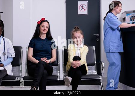 Smiling mother and daughter in busy private clinic waiting for pediatric doctor to do routine checkup while nurse is talking with receptionist. Woman and her child attending pediatrician appointment. Stock Photo